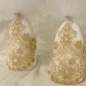 May include: Two white candle holders with gold lace designs. The candle holders are covered in a sheer white fabric.