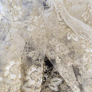 May include: A sheer, off-white tulle fabric featuring intricate, metallic gold floral embroidery. The design consists of detailed blossoms, swirling vines, and delicate leaves stitched throughout the translucent, lightweight mesh material.