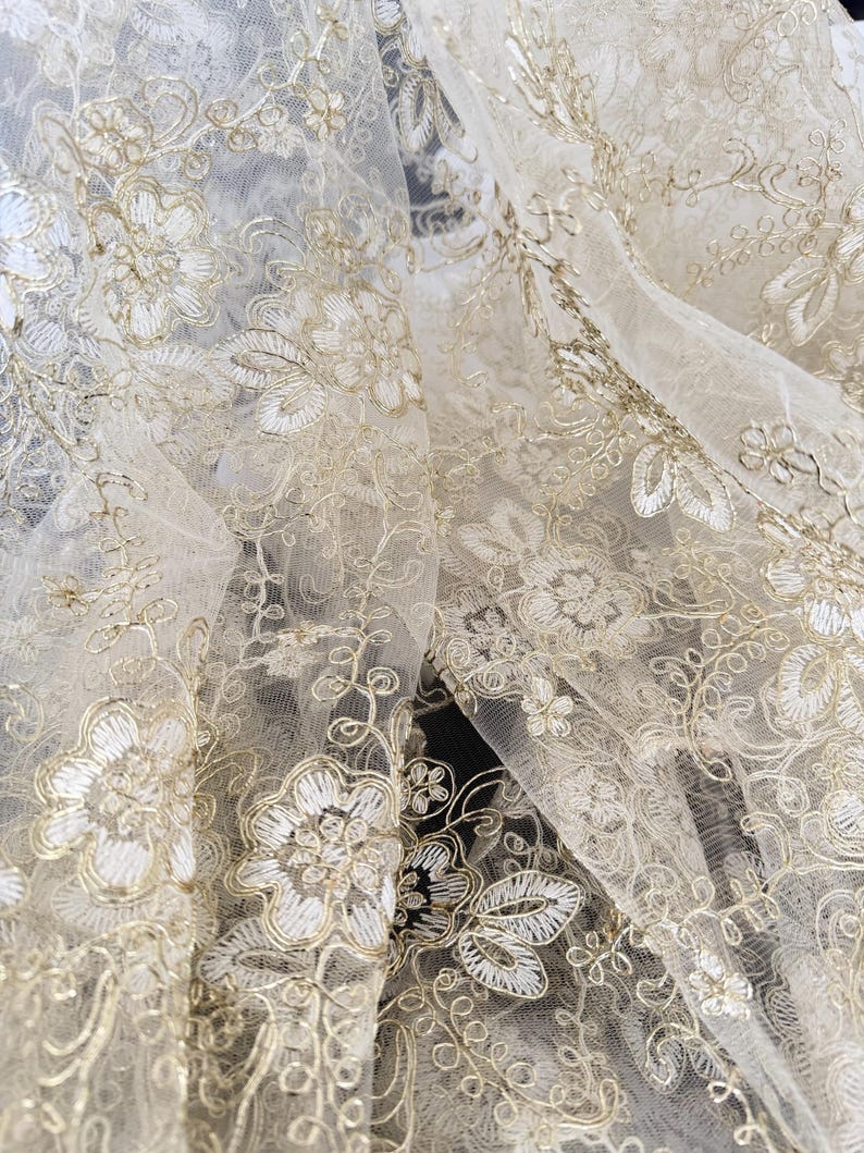 May include: A sheer, off-white tulle fabric featuring intricate, metallic gold floral embroidery. The design consists of detailed blossoms, swirling vines, and delicate leaves stitched throughout the translucent, lightweight mesh material.