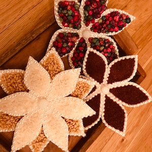Può includere: Tre vassoi decorativi a forma di fiore riempiti con diversi cereali e semi. I vassoi sono realizzati in materiale bianco e hanno un bordo perlato. I cereali e i semi sono disposti in uno schema simmetrico.