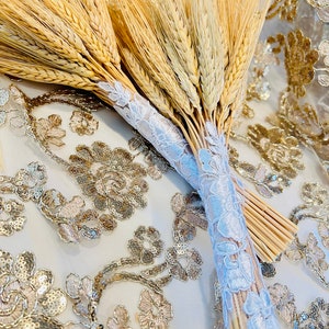 May include: A bouquet of dried wheat stalks tied together with a white lace ribbon. The bouquet is lying on a white fabric with gold sequins.