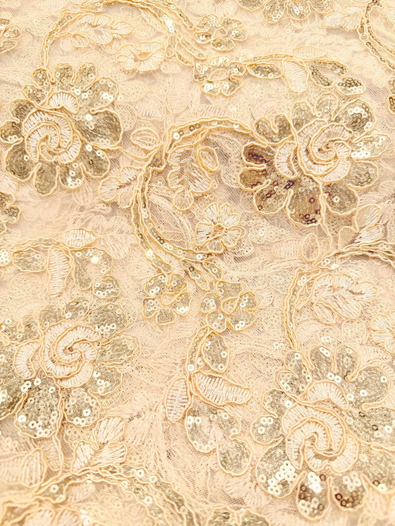 May include: A light beige lace fabric featuring an intricate floral pattern embroidered with matching thread and embellished with small, shimmering gold sequins throughout the petals and leaves, creating a textured and reflective surface design.