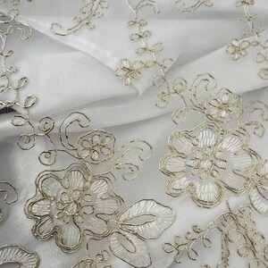 May include: A white sheer fabric featuring intricate silver metallic embroidery in a floral pattern. The design consists of detailed flower blossoms, swirling vines, and leaves, all outlined with a shimmering silver thread on a translucent base.
