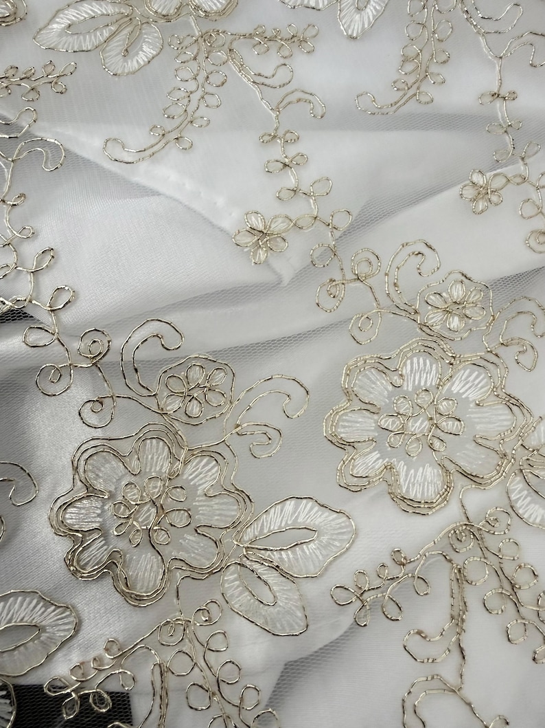 May include: A white sheer fabric featuring intricate silver metallic embroidery in a floral pattern. The design consists of detailed flower blossoms, swirling vines, and leaves, all outlined with a shimmering silver thread on a translucent base.