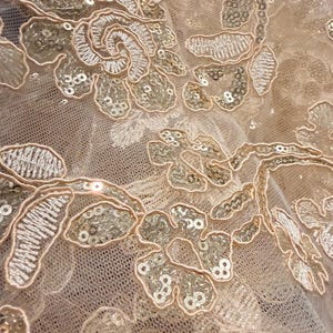 May include: A sheer beige mesh fabric featuring intricate floral embroidery in a matching thread color. The design is embellished with small, reflective sequins that follow the contours of the flower petals and leaves, creating a textured surface.