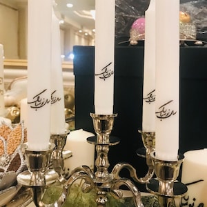 May include: A silver candelabra with five white candles. The candles are decorated with black calligraphy that reads "مُنْتَظِرُكَ".