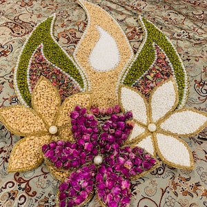 May include: A decorative floral arrangement featuring various grains, seeds, and dried flowers. The design includes green, beige, and white leaf-shaped elements, along with gold-trimmed flower shapes filled with pistachios and rice. The arrangement is adorned with pearls and dried rose buds.