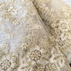 May include: A sheer fabric overlay featuring intricate gold metallic thread embroidery in a floral pattern. The design consists of detailed flower blossoms, swirling vines, and delicate leaf motifs stitched onto a fine, translucent mesh base.