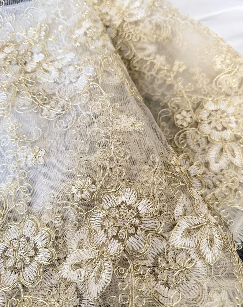 May include: A sheer fabric overlay featuring intricate gold metallic thread embroidery in a floral pattern. The design consists of detailed flower blossoms, swirling vines, and delicate leaf motifs stitched onto a fine, translucent mesh base.