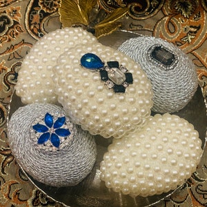 Puede incluir: Cinco huevos decorativos cubiertos de perlas, dos con detalles azules y plateados, dos con detalles plateados y uno con un detalle de joyas en blanco y negro.
