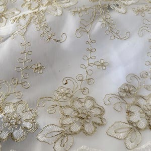 May include: A sheer white fabric featuring intricate gold metallic thread embroidery in a floral pattern. The design consists of detailed flowers, swirling vines, and small leaves, finished with a delicate scalloped edge along the bottom border.