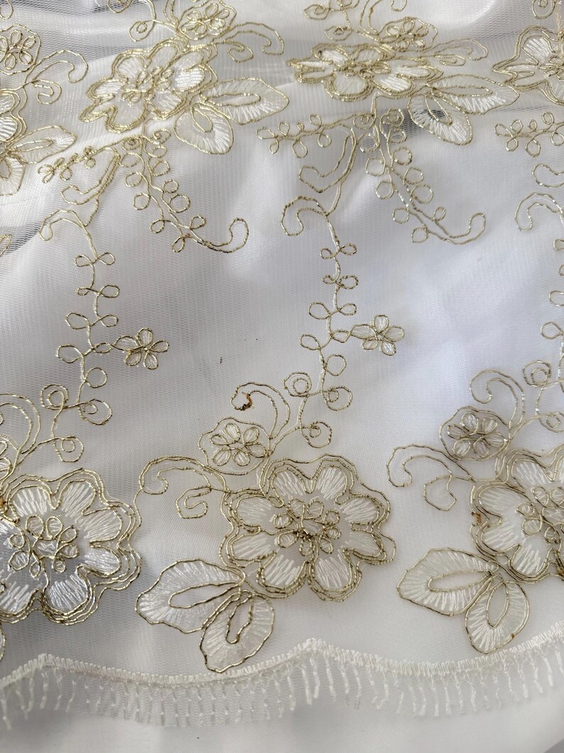 May include: A sheer white fabric featuring intricate gold metallic thread embroidery in a floral pattern. The design consists of detailed flowers, swirling vines, and small leaves, finished with a delicate scalloped edge along the bottom border.