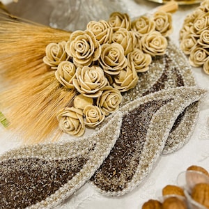 May include: A decorative arrangement of beige fabric roses on a white background. The roses are arranged in a bouquet shape and are surrounded by wheat stalks. The roses are on top of a beaded, brown and white, leaf-shaped tray.