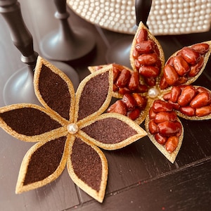 May include: Two decorative flower arrangements made with dried dates. One flower is brown with gold trim and a pearl in the center. The other flower is made with red dates and has a pearl in the center.