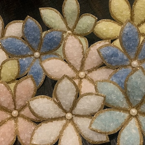 May include: A collection of six handmade paper flowers with a sparkly, glittery finish. Each flower has a different color: light blue, light pink, light green, and yellow. The flowers have a gold trim around the edges and a pearl in the center.