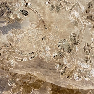 May include: A sheer champagne-colored mesh fabric featuring intricate floral embroidery with matching thread and scattered metallic sequins. The delicate netting base is adorned with repeating botanical patterns that create a textured surface.