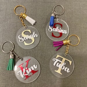 May include: Four clear acrylic keychains with different names and coloured tassels. The names are "Sarah", "Sakina", "Kim", and "Tuba". The tassels are white, blue, purple, and green.