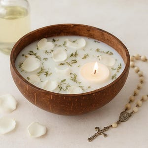 May include: A lit candle floats in a coconut bowl filled with milky liquid, white petals, and herbs. A rosary with a cross and a bottle of oil are also present. The bowl is brown, and the background is white.