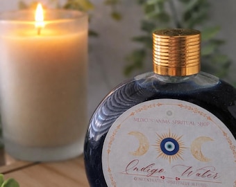 May include: A round, dark blue glass bottle with a gold cap, labeled "Indigo Water." The label includes text and a design with moons and an eye. A lit candle and greenery are in the background. The bottle contains 105 ml of liquid.