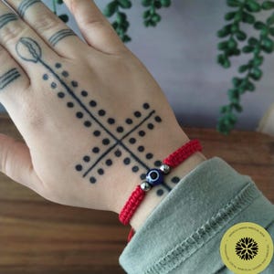 May include: Red string bracelet with a blue evil eye charm and silver beads. The bracelet is on a person's wrist.