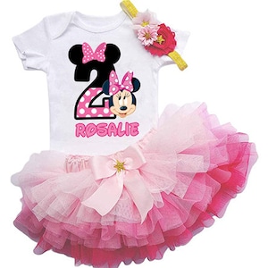 May include: A pink and white tutu with a white short-sleeved shirt featuring a pink and black Minnie Mouse graphic and the number 2. The shirt also has the name "Rosalie" printed on it. The outfit is decorated with a pink flower headband.