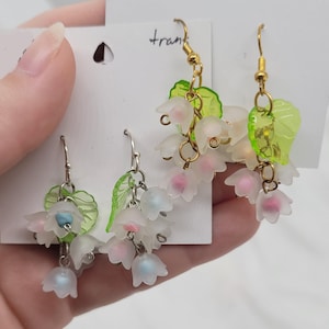 May include: Two pairs of dangle earrings. Each earring features clusters of white, pink, and blue flower-shaped beads with green leaf accents. The earrings have gold-coloured and silver-coloured hooks.