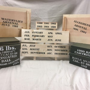 Puede incluir: Cajas y letreros de madera con texto negro con plantilla. Las cajas dicen "WATERVLIET ARSENAL JULY 1863" y "ALLEGHENY ARSENAL NOV. 1861." Los letreros muestran meses y años de 1860-1865. También dos cajas verdes con texto.