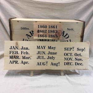 Puede incluir: Carteles de madera con texto negro. Un cartel enumera los meses del año y otro los años 1860-1865. Una caja de madera con el texto "50lbs" y "Arrowhead Mechanical" está en el fondo.
