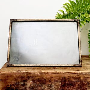 May include: A rectangular picture frame with a metallic bronze finish and a textured, speckled surface. The frame features decorative corner accents with intricate detailing and a clear glass front panel, providing a clean display area.