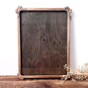 May include: A rectangular picture frame with a metallic copper-toned finish and ornate corner detailing. The frame features a dark brown wood-grain backing panel with visible natural wood textures and small dark hardware accents throughout the surface.