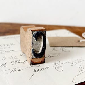 May include: A vintage letterpress printing block featuring the letter "C". The block has a wooden handle and a metal letter form. The letter is black and set against a gray background. The block rests on a sheet of paper with handwritten script.