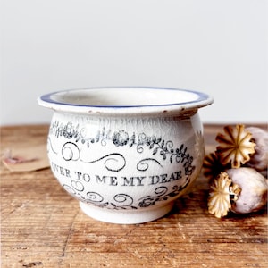 May include: A white ceramic bowl with a blue rim and a crackled glaze finish. The exterior features a black floral garland design and the printed text "OVER TO ME MY DEAR" in a serif font. The bowl has a rounded body and a flared top edge.