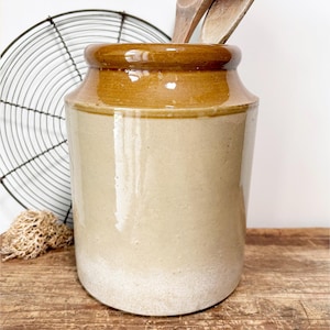 May include: A cylindrical stoneware crock featuring a two-tone glaze with a glossy dark brown rim and a smooth light beige body. The vessel has a wide mouth and a thick, rounded lip, providing a sturdy container for kitchen utensils or storage.