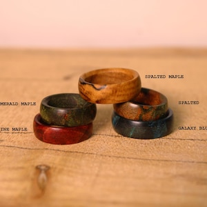 May include: A collection of five wooden rings with varied finishes, labeled as Wine Maple, Emerald Maple, Spalted Maple, Spalted, and Galaxy Blue Maple. The rings are stacked and arranged in a cluster, showcasing distinct wood grain patterns and colors.