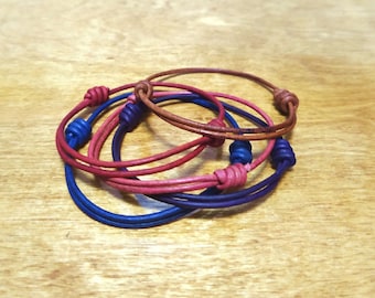 May include: Set of four leather bracelets in shades of brown, pink, and blue. Each bracelet has a knotted design and a small bead at the knot.