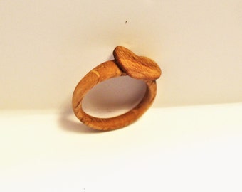 May include: A wooden ring with a heart-shaped top. The ring is made of light brown wood with a natural grain pattern.