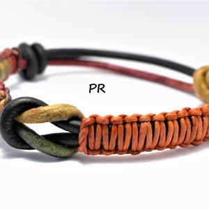 Puede incluir: Un brazalete de cuero multicolor con un diseño tejido y nudos. El brazalete está hecho de cuero negro, rojo, verde y marrón.