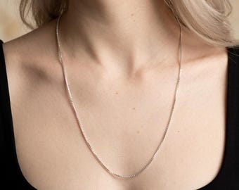 Könnte beinhalten: Eine silberne Box-Kettenhalskette. Die Halskette wird vor einer Person mit einem schwarzen Oberteil gezeigt. Die Kette ist zart und hängt über der Brust. Der Text "Hello Aura" befindet sich am unteren Rand des Bildes.