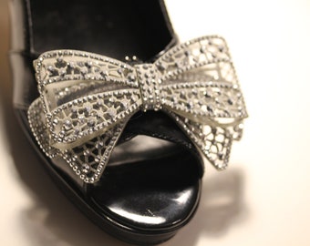 May include: Black open-toe sandal with a decorative bow. The bow is silver-colored, with a filigree design and embellished with small, sparkling rhinestones. The sandal has a glossy finish.