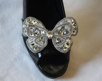 May include: Black patent leather peep-toe shoe with a sparkling bow embellishment. The bow is adorned with numerous clear, faceted crystals and small silver beads, creating a glamorous accessory.