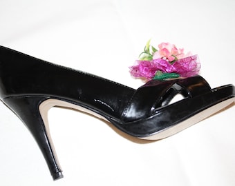 May include: A black patent leather peep-toe platform heel with a stiletto heel. A decorative floral arrangement of pink and purple flowers adorns the top of the shoe. The shoe is set against a white background.