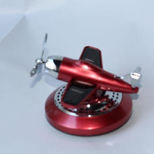 Puede incluir: Un objeto decorativo con forma de avión rojo y plateado. El avión tiene una hélice y se apoya sobre una base circular. La base y el avión son de color rojo brillante, con detalles plateados. Probablemente sea un ambientador para coche o una pieza decorativa.