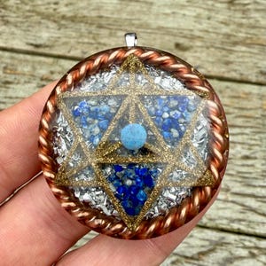 Puede incluir: Colgante redondo con diseño de Estrella de David, detalles dorados, piedras preciosas azules y una esfera azul claro. El colgante está encapsulado en resina transparente con un borde de cuerda color cobre.