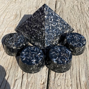 May include: A collection of black resin objects, including a pyramid and five round, flat-topped pieces. The items have a textured, speckled appearance with silver and black tones. The objects are arranged on a weathered wooden surface.