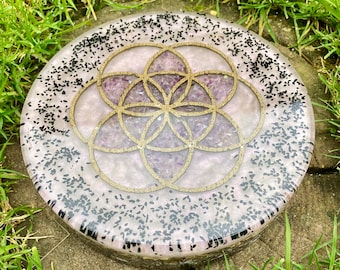 May include: A circular resin coaster featuring a gold-colored geometric flower of life pattern embedded in a light purple base with small purple crystal fragments and scattered black flecks throughout the translucent material.