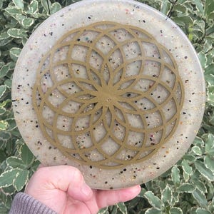 Puede incluir: Posavasos redondo de resina color crema con un diseño geométrico dorado. El posavasos está salpicado de pequeñas piedras de colores y tiene un patrón circular dorado en capas en el centro. El posavasos se sostiene en una mano.