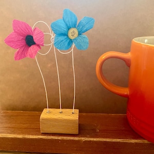 Op de afbeelding: Een decoratieve presentatie met twee papieren bloemen, één roze en één blauw, op een houten voet. De roze bloem heeft een zwart hart, de blauwe een geel hart. Een oranje mok staat op de achtergrond.