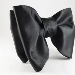 May include: A large, black satin bow tie. The bow tie is made of a shiny, smooth fabric and is tied in a classic bow shape. The bow tie is a formal accessory, suitable for special occasions.