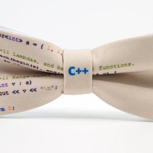 K&ouml;nnte beinhalten: Ein beiger Fliege mit einem Muster aus C++-Code darauf gedruckt. Die Fliege hat ein blaues C++-Logo in der Mitte.