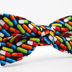 May include: A black bow tie with a colorful pattern of pills. The pills are red, orange, yellow, green, and blue.